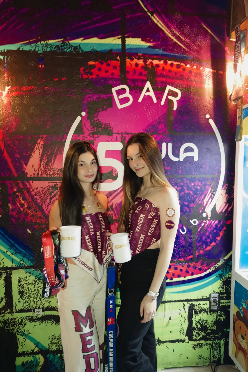 Duas integrantes da atlética com canecas e tops personalizados no bar