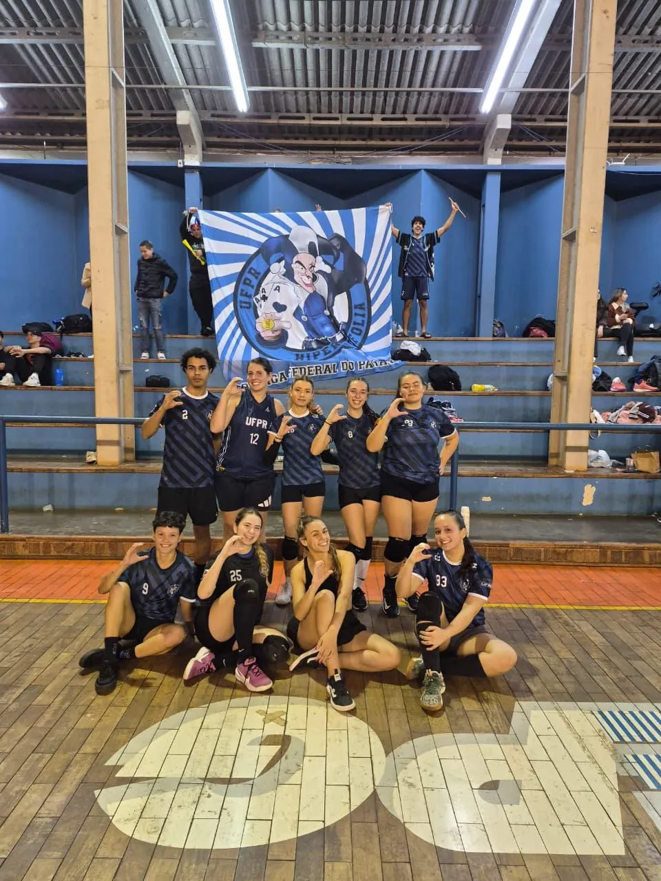 Time de vôlei da atlética posando diante da bandeira no ginásio
